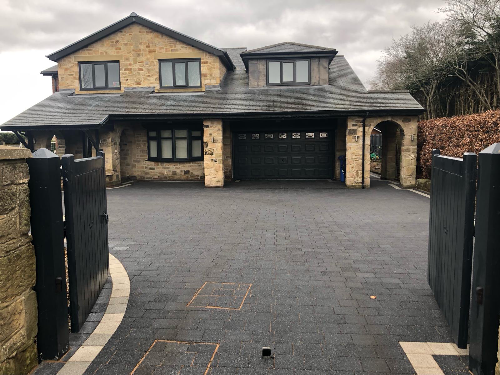 Charcoal block paved driveway between timber gates running to a double garage, with recessed covers set into the pattern — The Vale Company