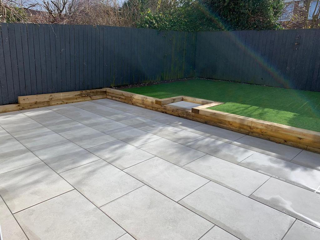 Light grey porcelain terrace with a timber sleeper retaining edge holding back a raised lawn — The Vale Company