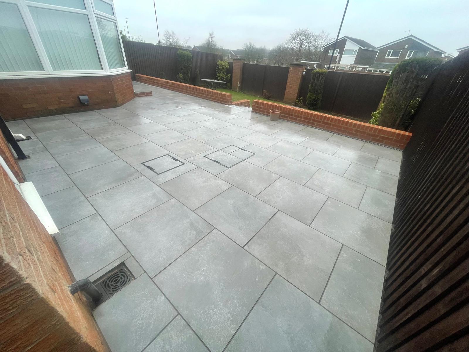 Wide grey porcelain terrace bounded by a brick retaining wall and channel drainage — The Vale Company