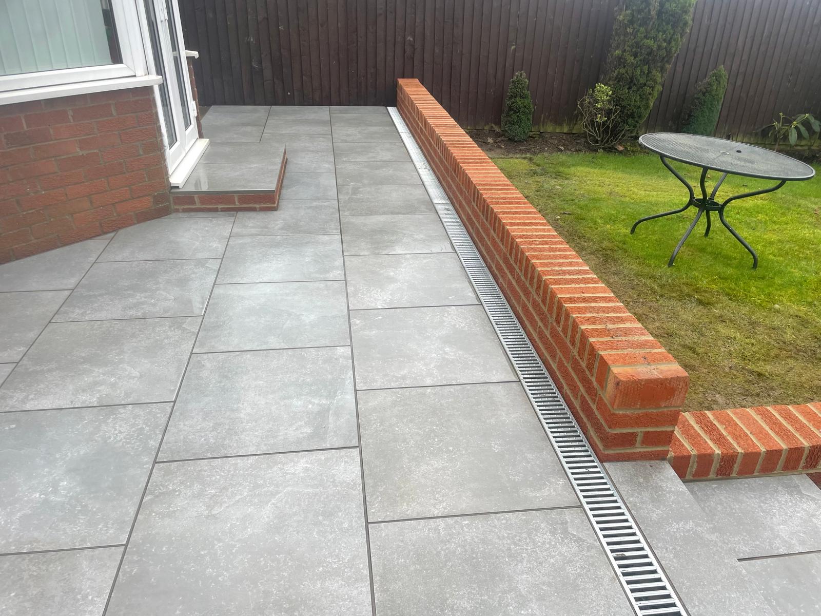 Grey porcelain terrace with a linear channel drain running along a brick retaining wall — The Vale Company