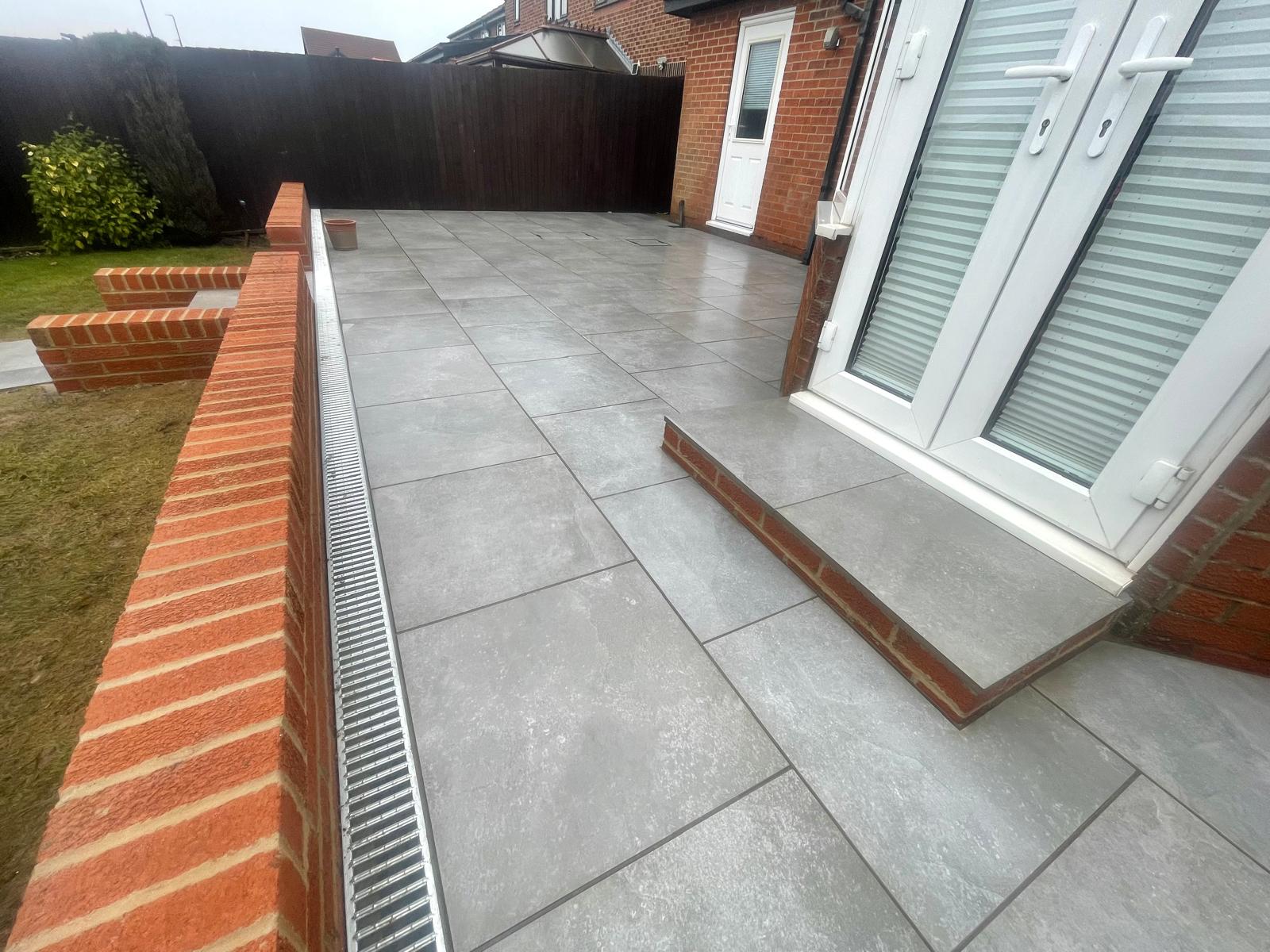 Wide view of a grey porcelain terrace with a channel drain against a brick retaining wall — The Vale Company