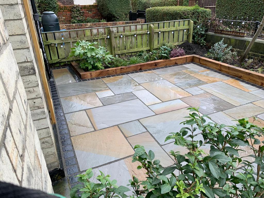 Grey and buff Indian sandstone patio with a charcoal sett border and timber sleeper planting beds — The Vale Company